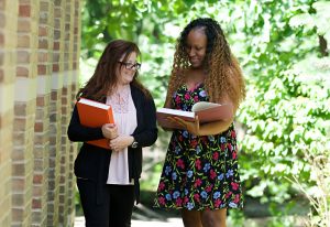 Photo of two women walking outside and conversing while looking at a books. 