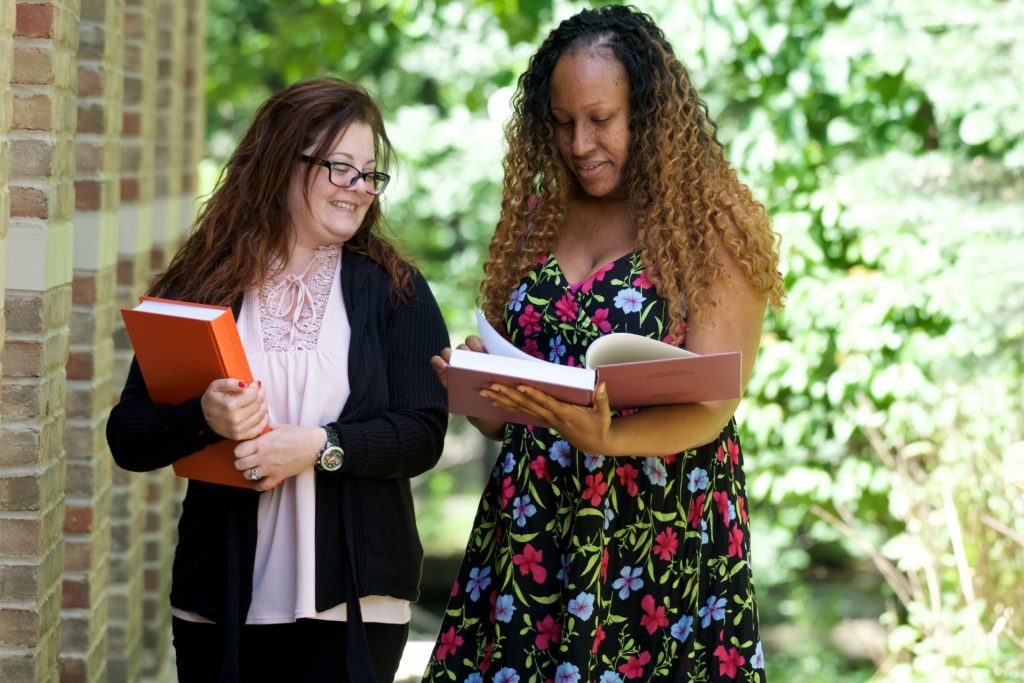 Photo of two women standing together outside next to a brick wall with trees in the background. One woman has a book open ans is looking at it. The other woman is holding a book in her arms and smiling. She is looking over at the woman holding the book open.