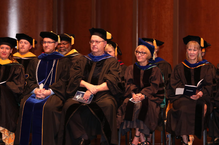 Photo of MSP faculty in graduation regalia sitting on a stage during graduation.