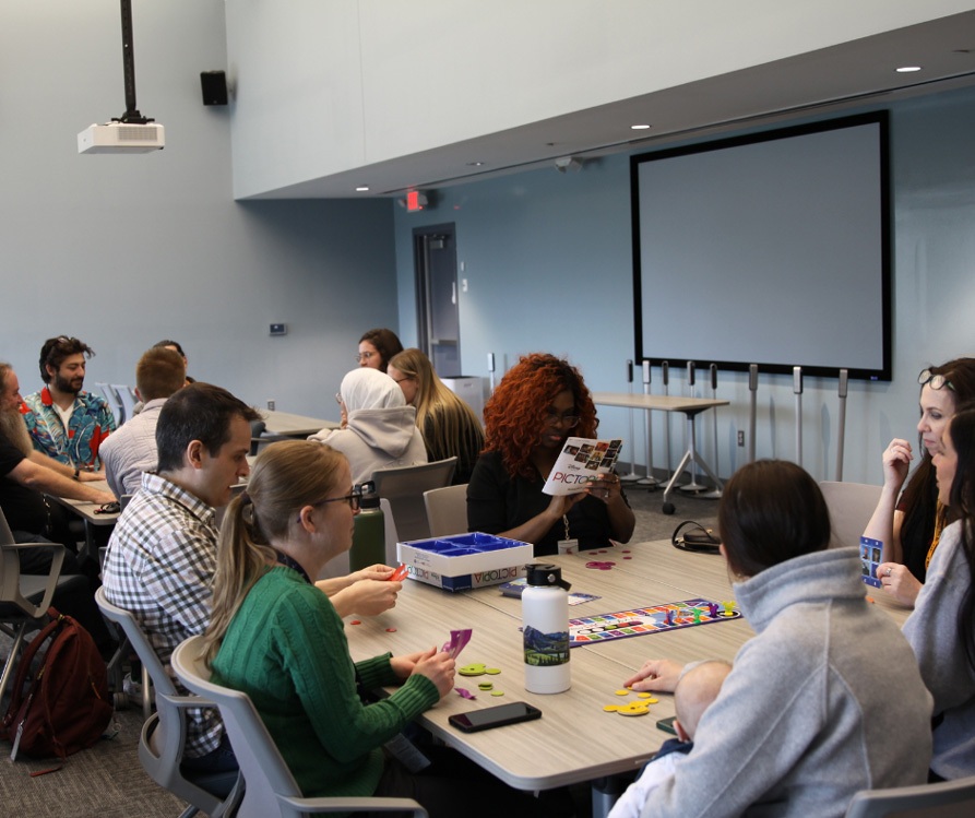 Photo of students, faculty, and staff sitting at a table playing Disney Pictopia with another table behind.