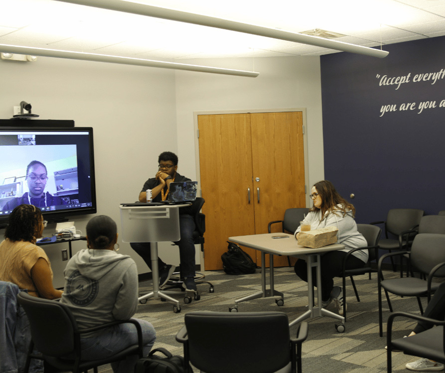 Photo of 4 students sitting in a semi circle with one more on screen having joined from zoom.