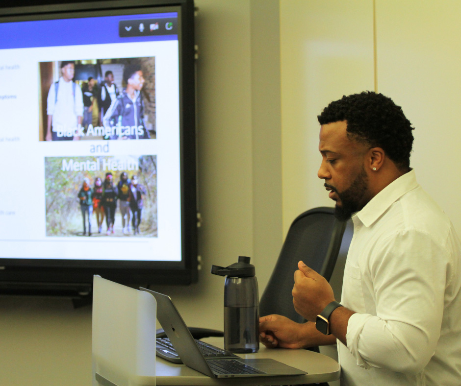 Ed-Dee Williams standing at a classroom podium with laptop with a presentation labeled "Black Americans and Mental Health" in the background.