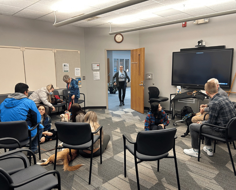 Wide shot photo of students sitting in chairs and on the floor across a classroom petting therapy dogs.