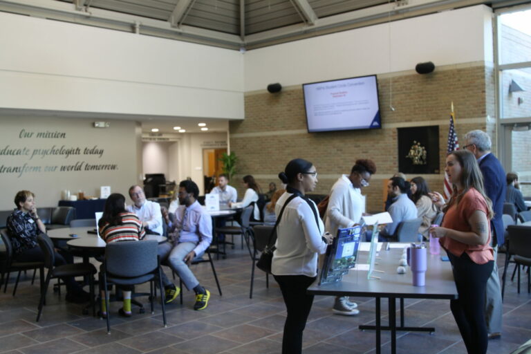 Photo from Open House featuring some staff and prospective students standing at a display table talking with others sitting at smaller tables in the background.