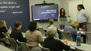 Photo of presenters at the Cultural Competency Conference in 2023 standing at the front of a room to the side of the screen. Audience is sitting in chairs.