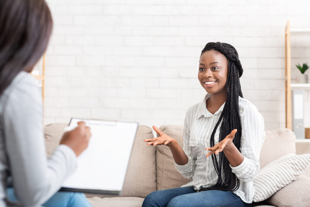 Stockphoto of two women in a therapy session. One has a notebook and is writing in it and the other is sitting across from her smiling and talking.