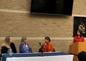 Three female panelists sitting at a table with one holding a microphone. Carrie standing to the side at a podium.