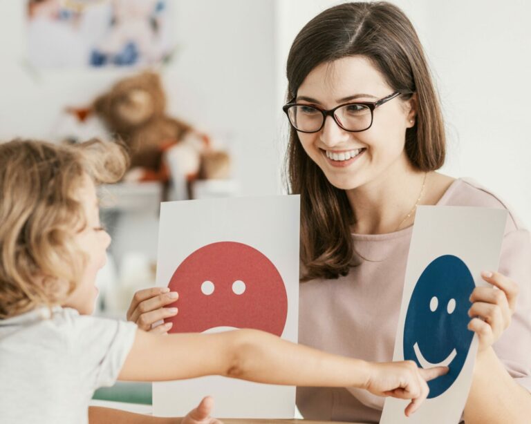 Woman therapist holding up a picture of a smiling face in one hand and a frowning face in the other. Child smiling and pointing at the smiling face.