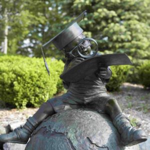 Bronze statue of child sitting on a globe wearing a mortar board holding a book up to their face with a magnifying glass in hand looking at a page