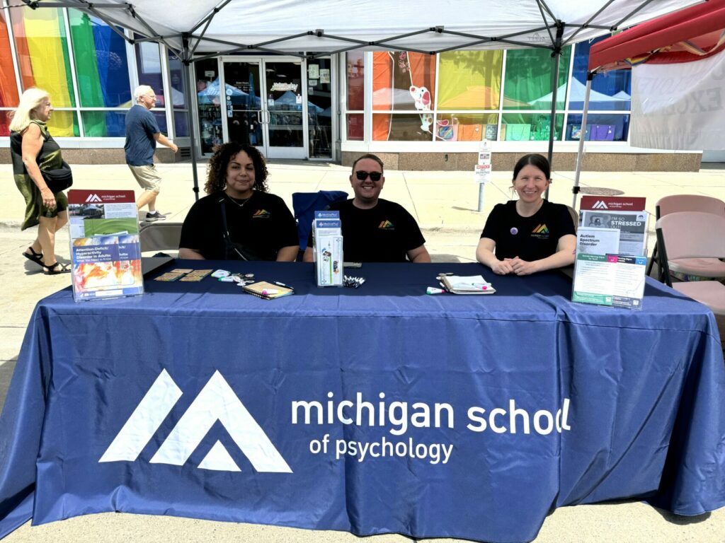 Photo of two students and staff sitting at a table with information materials, windows decorated with the pride flag serve as the background.