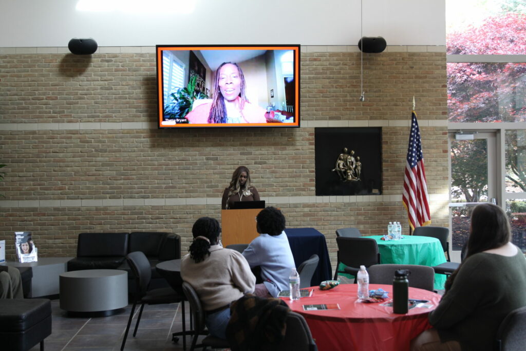 Students sitting at tables in the atrium with one student at a podium and a guest speaker on zoom above.