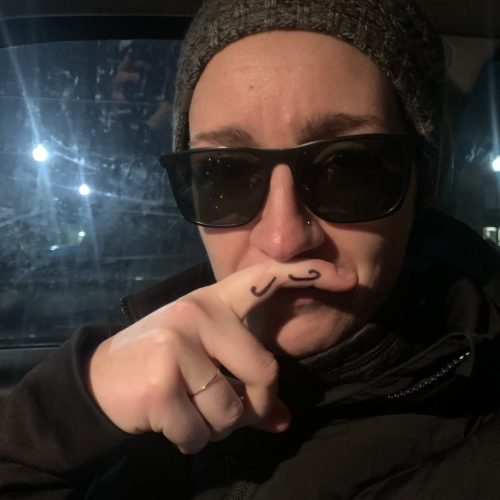 Photo of a student wearing a beanie and sunglasses holding up a finger with a drawn on mustache to their mouth